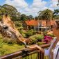 A smiling traveler feeds a Rothschild giraffe from the Giraffe Centre viewing platform in Nairobi, with the beautiful Karen Blixen Museum, colorful gardens, and lush greenery visible in the background during a guided tour by Cruzeiro Safaris.